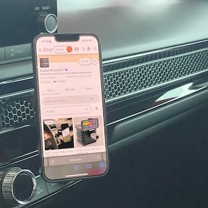 Op de afbeelding: Een smartphone met een Etsy-winkel is gemonteerd op het dashboard van een auto. De telefoon wordt vastgehouden door een magnetische houder. Het dashboard is zwart met zilveren accenten en een honingraatvormig ventilatierooster.