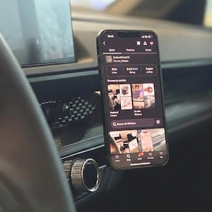 以下が含まれることがあります： 車のダッシュボードに取り付けられたスマートフォン。eコマースアプリが表示されています。黒いスマートフォンには、製品画像が表示されたストアフロントが表示されています。車のダッシュボードとステアリングホイールが背景に見えます。