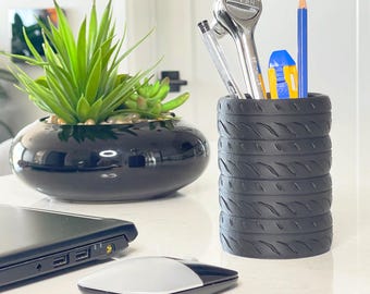 May include: A black tire-shaped desk organizer filled with writing utensils, including a pencil, eraser, and a silver ratchet. A black ceramic planter with a green succulent sits to the left. A laptop and mouse are in the foreground.