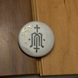 Puede incluir: Un botón blanco con un gráfico en blanco y negro de una cruz estilizada y la letra M. El botón está sobre una superficie de madera.