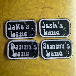 May include: Four black and white embroidered patches with white text and yellow accents. The patches read "Jake's Lane", "Josh's Lane", "Danny's Lane", and "Sammy's Lane".