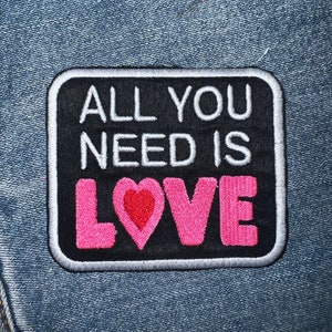 May include: A black and white embroidered patch with a white border. The patch says "All You Need Is Love" in white text with a pink heart in the middle of the word "Love".