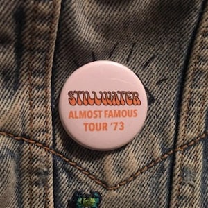 May include: A round, pink button with the text "STILLWATER" and "ALMOST FAMOUS TOUR '73" in orange lettering. The button is pinned to a denim jacket.