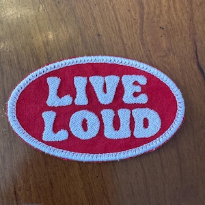 Puede incluir: Un parche ovalado rojo con texto bordado blanco "LIVE LOUD". El parche tiene un borde cosido blanco. El parche está sobre una superficie de madera.