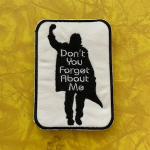 Könnte beinhalten: Weißer gestickter Aufnäher mit einer schwarzen Silhouette einer Person, die die Faust in die Luft reckt. Der Text "Don't You Forget About Me" steht unter der Silhouette.