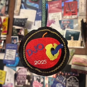 以下が含まれることがあります： 赤いリンゴのデザイン、黄色のミミズ、そして年号「2025」の上に「DJO」の文字が刺繍された丸いオーナメント。 オーナメントは、金色のステッチが施された黒い縁取りと、吊り下げ用の緑と黒のリボンが付いています。