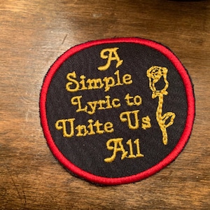 May include: A black and red circular patch with gold embroidery. The text reads "A Simple Lyric to Unite Us All" with a stylized rose design.