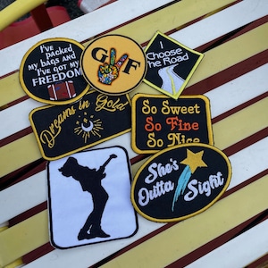 May include: A collection of seven embroidered iron-on patches with various designs and text. The patches are on a yellow and white striped surface. The patches include a peace sign with the letters "GF" inside, a road with the text "I Choose the Road", a black patch with the text "Dreams in Gold", a black patch with the text "So Sweet So Fine So Nice", a black patch with a silhouette of a person playing guitar, a black patch with the text "She's Outta Sight" and a yellow patch with the text "I've packed my bags and I've got my freedom".