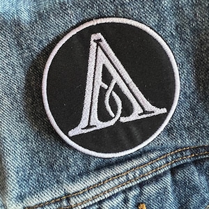Può includere: Una toppa ricamata in bianco e nero con una lettera "A" stilizzata in un cerchio. La toppa è cucita su una giacca di jeans blu.
