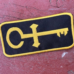 Peut inclure: Un écusson brodé noir et jaune avec un motif de clé et de cercle. La clé est stylisée avec une poignée en forme de croix.