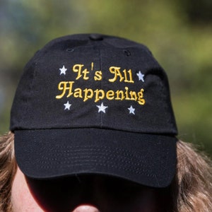 May include: Black baseball cap with white stars and the text "It's All Happening" in yellow embroidery.