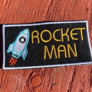May include: A rectangular black patch with the words "ROCKET MAN" embroidered in yellow. A light blue rocket with orange flames is on the left side. The patch has a white border.