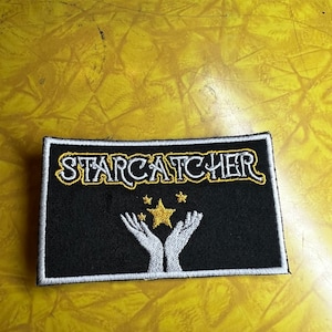 May include: Black embroidered patch with white border and gold stitching. The patch features the text "STARCATCHER" and a design of two hands reaching up to a star.