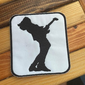 May include: Black and white embroidered patch of a person playing a guitar. The patch is rectangular and has a white background.