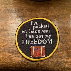 Pode incluir: Um remendo redondo bordado em preto e dourado com o texto "I've packed my bags and I've got my FREEDOM" e uma pequena mala marrom com detalhes vermelhos e azuis.