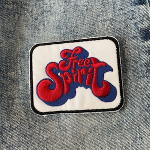 May include: A white patch with black edging features a red and blue embroidered design that reads "Free Spirit".