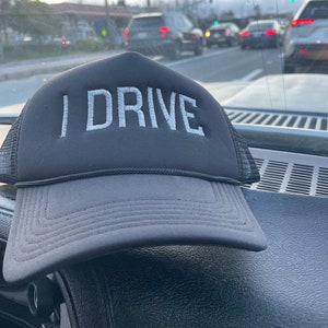 May include: A black trucker hat with the white embroidered text "I DRIVE". The hat has a mesh back and a curved brim. The hat is resting on a black dashboard, with a blurred background of a road and cars.