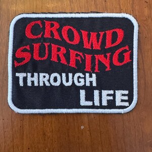 May include: Black rectangular patch with white border and the words "CROWD SURFING THROUGH LIFE" in red and white lettering. The patch is likely for a jacket or bag.