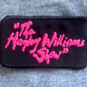 May include: A rectangular black embroidered patch with rounded corners featuring the text "The Hayley Williams Show" stitched in bright pink thread. The patch has a solid black border and is displayed against a textured blue denim fabric background.