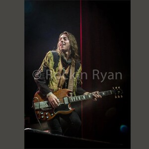 Peut inclure: Un homme aux cheveux longs et bruns joue de la guitare électrique brune avec des accents dorés sur le corps et un pickguard noir. Il porte une veste brune avec des franges dorées et un bandana noir. L'arrière-plan est sombre avec un projecteur rouge.