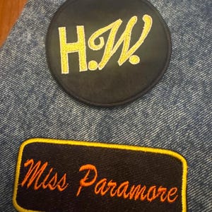 May include: Two embroidered patches on denim. One is a black circle with the yellow letters "H.W." The other is a black rectangle with the orange text "Miss Paramore" and a yellow border.