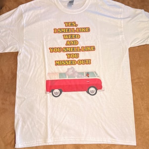 May include: White t-shirt featuring a graphic of a red Volkswagen van with a person driving and smoke coming out of the back. The text on the shirt reads "YES, I SMELL LIKE WEED AND YOU SMELL LIKE YOU MISSED OUT!"