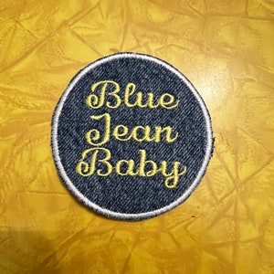 May include: A blue denim patch with white stitching around the edges. The text "Blue Jean Baby" is embroidered in yellow thread.