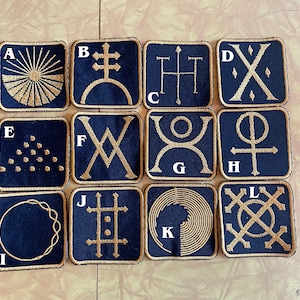 May include: Twelve square patches with gold embroidery on a dark blue background. Each patch has a letter from A to L and a different geometric design.