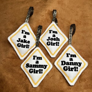 May include: Four white diamond-shaped ornaments with gold trim and black embroidery. Each ornament has a different name on it: "I'm a Jake Girl!", "I'm a Josh Girl!", "I'm a Sammy Girl!", and "I'm a Danny Girl!"