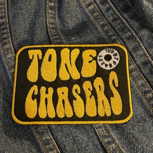 May include: A black and yellow embroidered patch with the text "TONE CHASERS" in a retro font. A small white circle with black numbers is in the upper right corner of the patch.