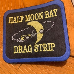 May include: A black embroidered patch with a blue border. It features a yellow crescent moon with a car driving around it. The text "HALF MOON BAY DRAG STRIP" is embroidered in yellow.