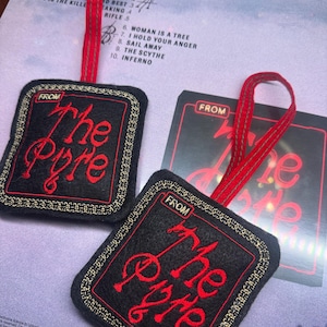 Puede incluir: Dos adornos de fieltro negro con texto bordado en rojo "The Pyre" y un borde dorado, sujetos a una cinta roja. Los adornos tienen forma cuadrada con esquinas redondeadas. También se ve una caja de CD.