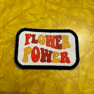 May include: A white embroidered patch with black trim featuring the text "FLOWER POWER" in a retro font with orange, yellow, and red lettering.