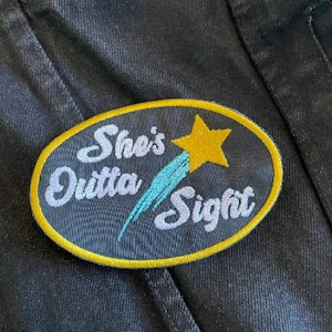 May include: A black and gold oval patch with the text "She's Outta Sight" in white cursive lettering. A blue shooting star with a yellow star at the end is behind the text.