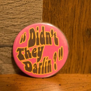 Può includere: Un bottone rosa con il testo "Didn't they darlin?" in lettere nere e gialle.