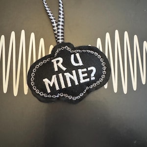 May include: A black felt ornament with the embroidered text "R U MINE?" in white. The ornament has a scalloped edge and a black and white checkered ribbon for hanging. The background is dark with white wave-like lines.