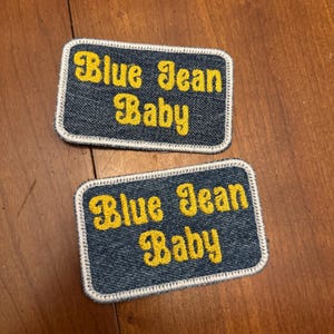 May include: Two denim patches with the text "Blue Jean Baby" in yellow script. The patches have a white border and are rectangular in shape. The patches are on a wooden surface.