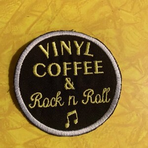 Puede incluir: Un parche bordado en blanco y negro con el texto "Vinyl Coffee & Rock n Roll" en hilo amarillo. Una nota musical está bordada debajo del texto.