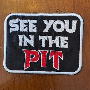 May include: Black rectangular patch with white embroidered border. The patch features the text "SEE YOU IN THE PIT" in white and red embroidered letters. The patch is likely for a jacket or bag.