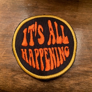 May include: A round, black patch with a gold border. The patch has orange embroidered text that reads "IT'S ALL HAPPENING".