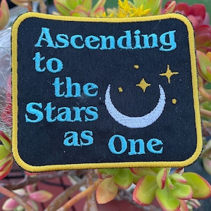 Può includere: Una toppa ricamata nera con cuciture gialle intorno ai bordi. La toppa presenta una luna crescente con stelle e il testo "Ascending to the Stars as One" in blu.