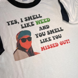 May include: White t-shirt with a black ringer neck. The shirt has a graphic of a person with sunglasses and the text "YES, I SMELL LIKE WEED AND YOU SMELL LIKE YOU MISSED OUT!"