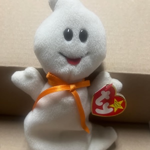 Può includere: Un peluche bianco a forma di fantasma beanie baby con una bocca rossa, occhi neri e un nastro arancione. Il beanie baby ha un'etichetta Ty rossa e bianca attaccata che dice "Beanie Babies" e ha una stella.