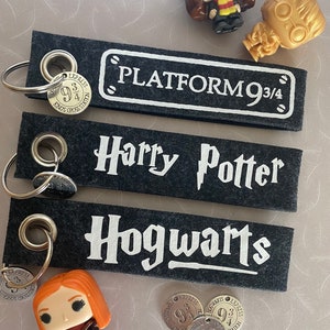 Könnte beinhalten: Drei schwarze Schlüsselanhänger aus Filz mit weißen Buchstaben. Die Schlüsselanhänger lauten "Platform 9 3/4", "Harry Potter" und "Hogwarts". Außerdem gibt es drei silberne Metallanhänger mit dem Text "Express 9 3/4 Kings Cross Station" und zwei kleine Plastikfiguren von Harry-Potter-Charakteren.