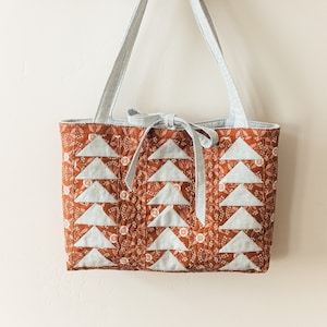 May include: A brown and white quilted tote bag with a floral pattern and gray straps. The bag is hanging on a wooden rack with hooks.