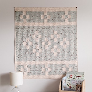 May include: A light blue and pink quilt with a checkered pattern hangs on a white wall. The quilt has a heart shape in the center. There are two books and a lamp on a white table in front of the quilt.