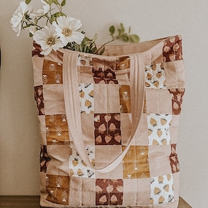 Könnte beinhalten: Eine gesteppte Tragetasche mit Patchwork-Design aus braunen, rosa und weißen Stoffquadraten mit Erdbeerprint. Die Tasche hat zwei lange Henkel und steht auf einer Holzoberfläche.