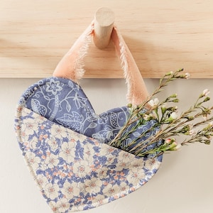 May include: A heart-shaped fabric pouch with a floral pattern hangs from a wooden hook. The pouch is filled with a bouquet of small white flowers.