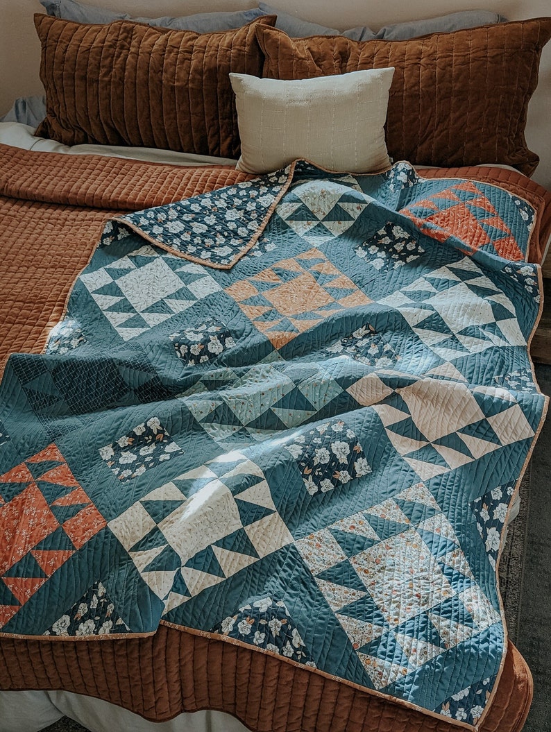 May include: A blue and white quilt with a geometric pattern. The quilt is made of different fabrics and has a brown border. The quilt is folded on a bed with brown pillows.