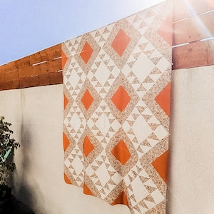 May include: A white and orange quilt with a geometric pattern. The quilt is hanging on a wall.